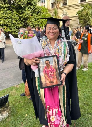 Tofilau standing in her graduation gown and hat. Holding flowers and a framed picture of her mother